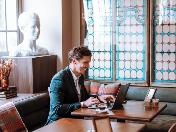 Un hombre trabajando en una computadora portátil sobre una mesa de madera, con una taza de café, en una habitación que tiene una escultura de busto y ventanas con diseños.