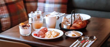 Uma mesa posta para o café da manhã, com croissant, queijo, frutas e cereal, acompanhada de uma xícara de café e um copo de suco de laranja.