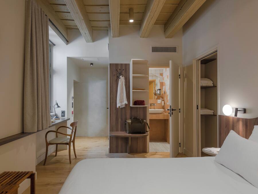Habitación de hotel con cama, escritorio, silla, armario y baño visible a través de una puerta abierta.