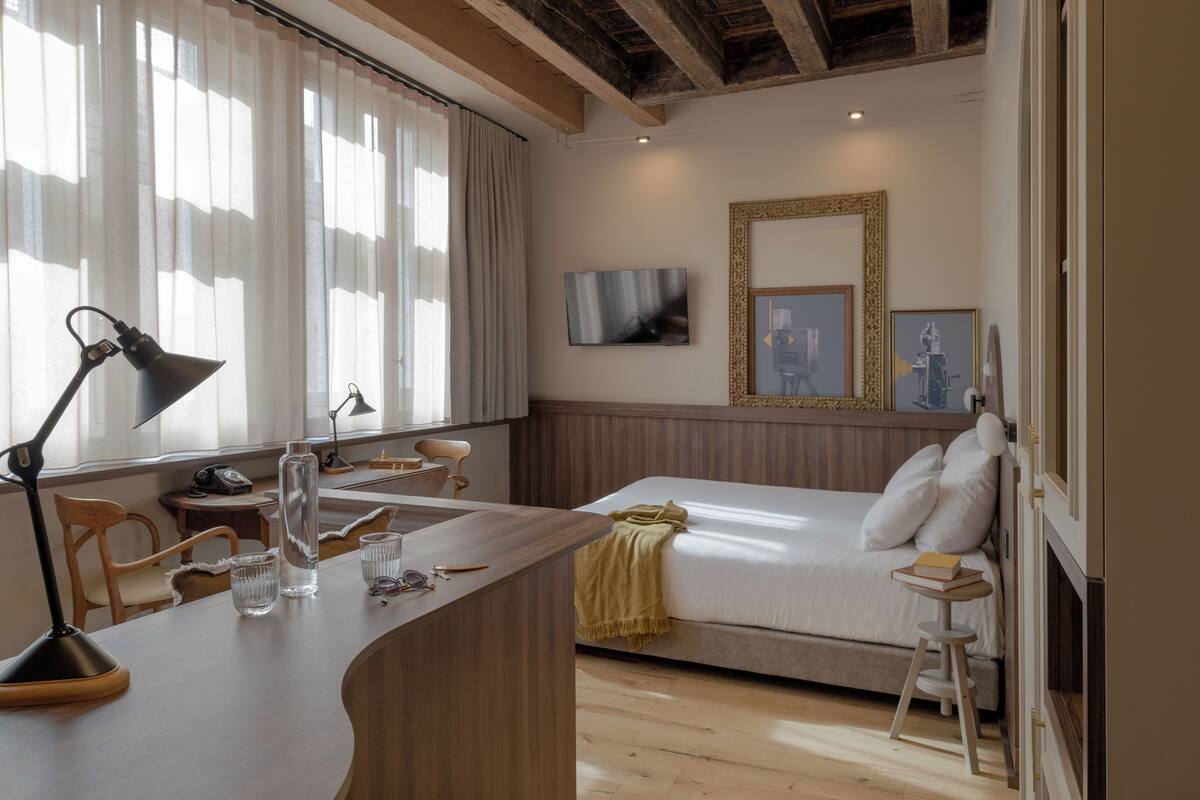 A hotel room with a bed, a desk with a lamp, a chair, a TV, and artwork on the wall.