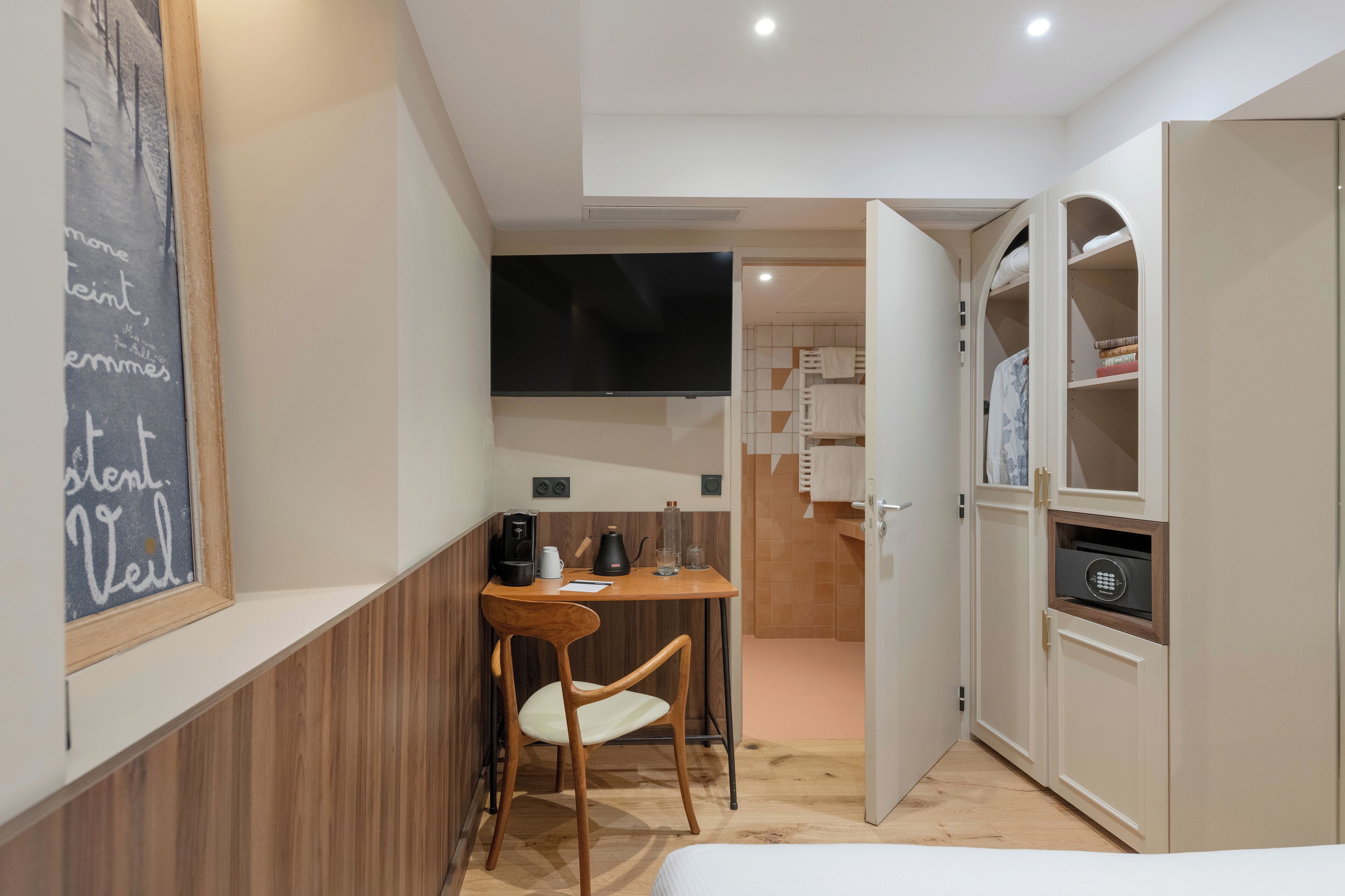 A compact hotel room with a wooden desk, a chair, a bed, a television, and built-in storage cabinets.