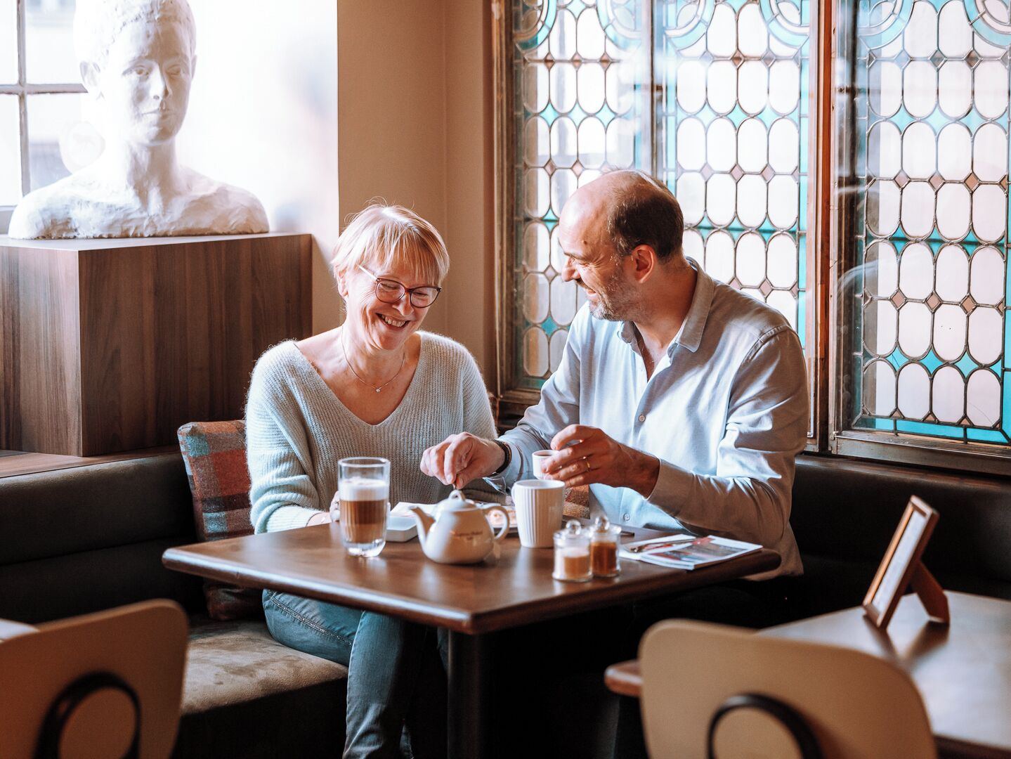 Twee personen zitten aan een tafel met een theepot en kopjes, in een kamer met een glas-in-loodraam en een buste.