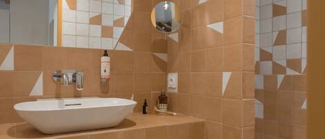 Un baño con un lavabo blanco, un espejo y una ducha. Hay un banco de madera con toallas debajo del lavabo.