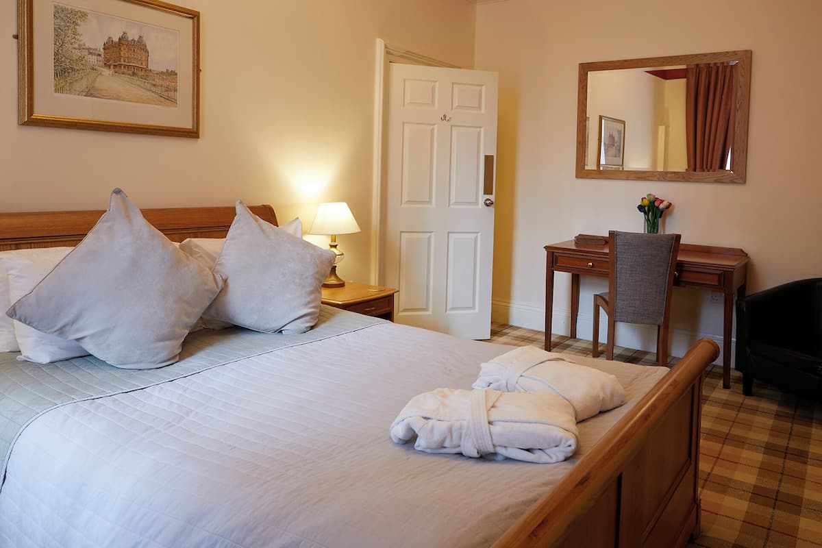A hotel room with a bed, pillows, a desk, a chair, a lamp, a mirror, and a painting on the wall.