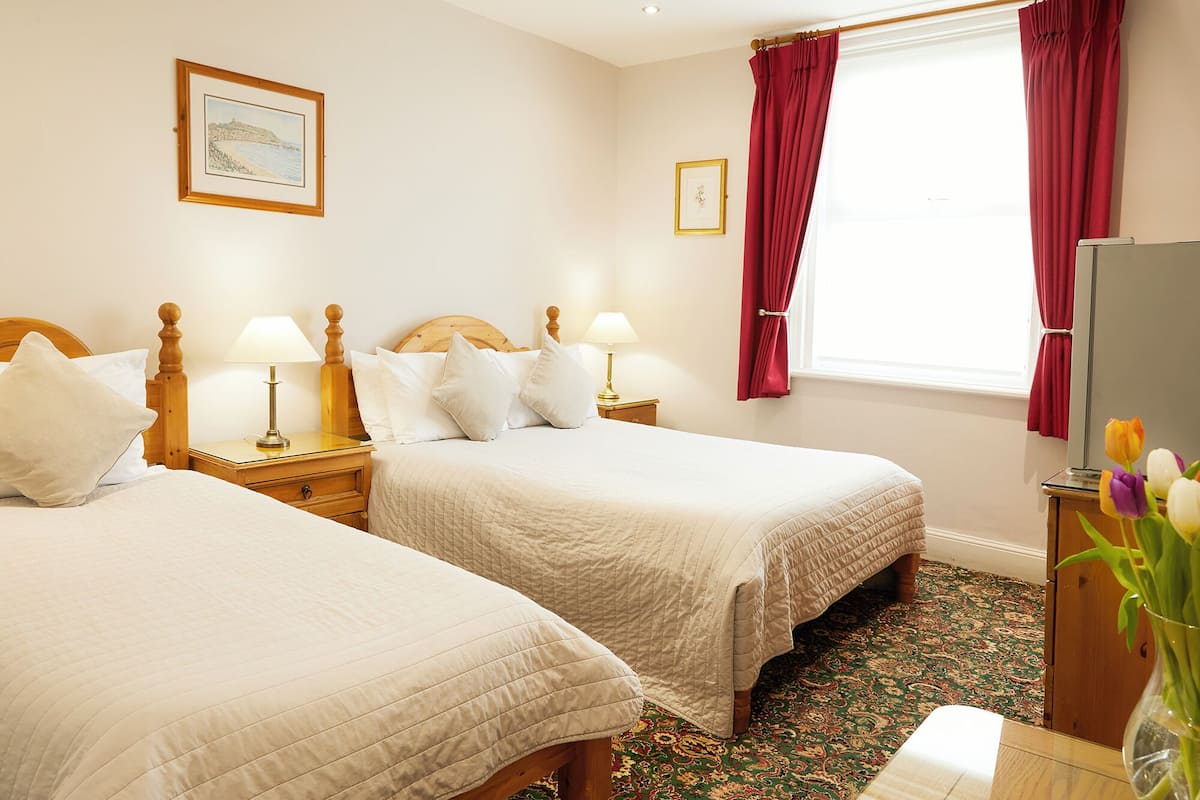 A hotel room with two beds, a wooden headboard, bedside lamps, a window with red curtains, and a floral arrangement.