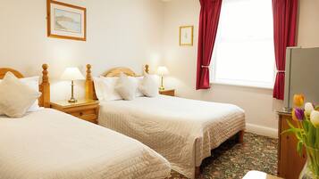 A hotel room with two beds, a wooden headboard, bedside lamps, a window with red curtains, and a floral arrangement.
