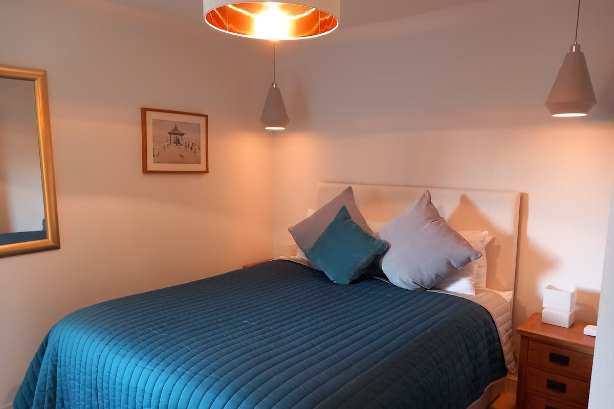 A neatly made bed with a blue quilt, a bedside table with a lamp, and a framed picture on the wall.