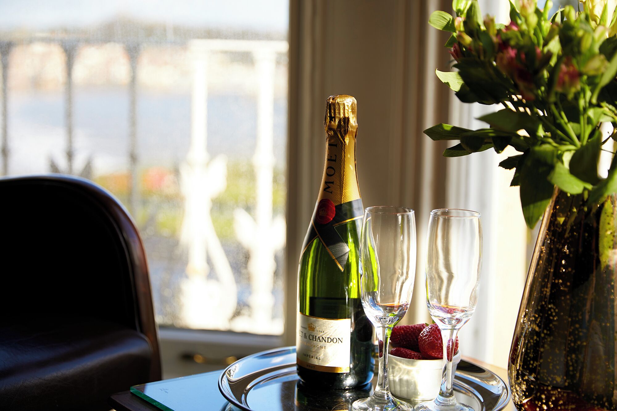 A bottle of Moët & Chandon champagne, two glasses, and a vase with flowers on a table by a window.