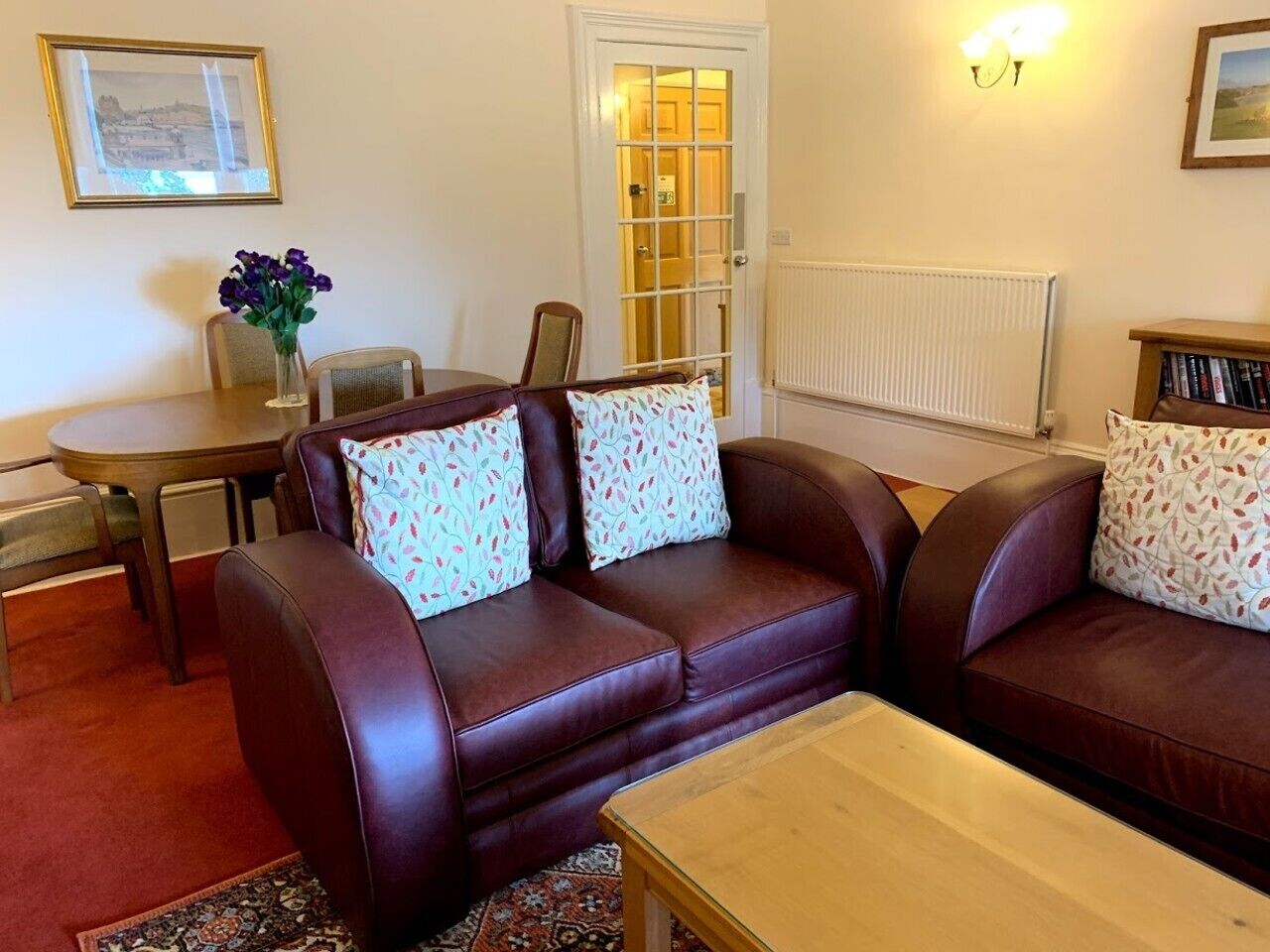 A living room with a brown leather sofa, a wooden coffee table, a dining table with chairs, and a radiator.