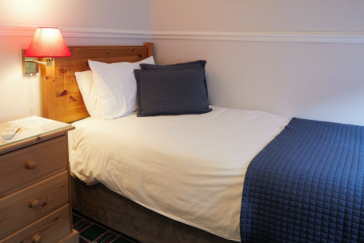A neatly made bed with white and dark blue bedding, a wooden headboard, a bedside lamp, and a wooden nightstand with drawers.