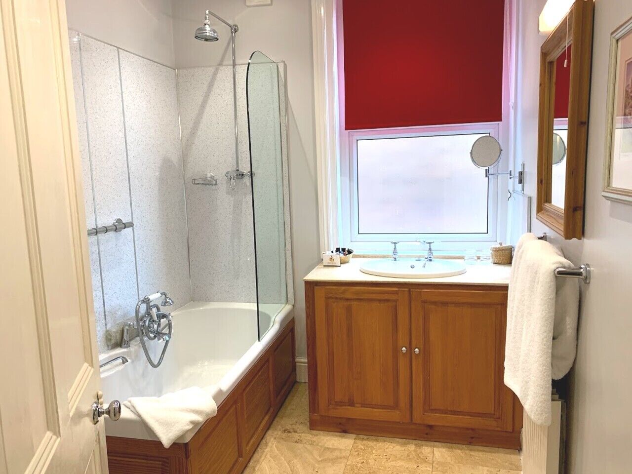 A bathroom with a glass-enclosed shower, a freestanding bathtub, wooden cabinets, a sink with a mirror, and a window with a red blind.