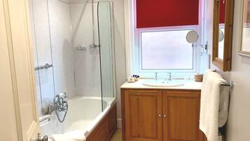 A bathroom with a glass-enclosed shower, a freestanding bathtub, wooden cabinets, a sink with a mirror, and a window with a red blind.
