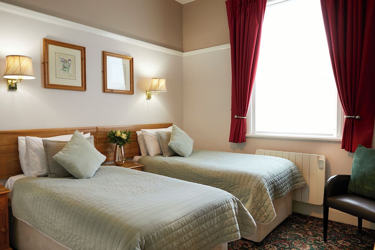 A hotel room with two beds, a nightstand, a chair, a window with red curtains, and wall-mounted lamps.