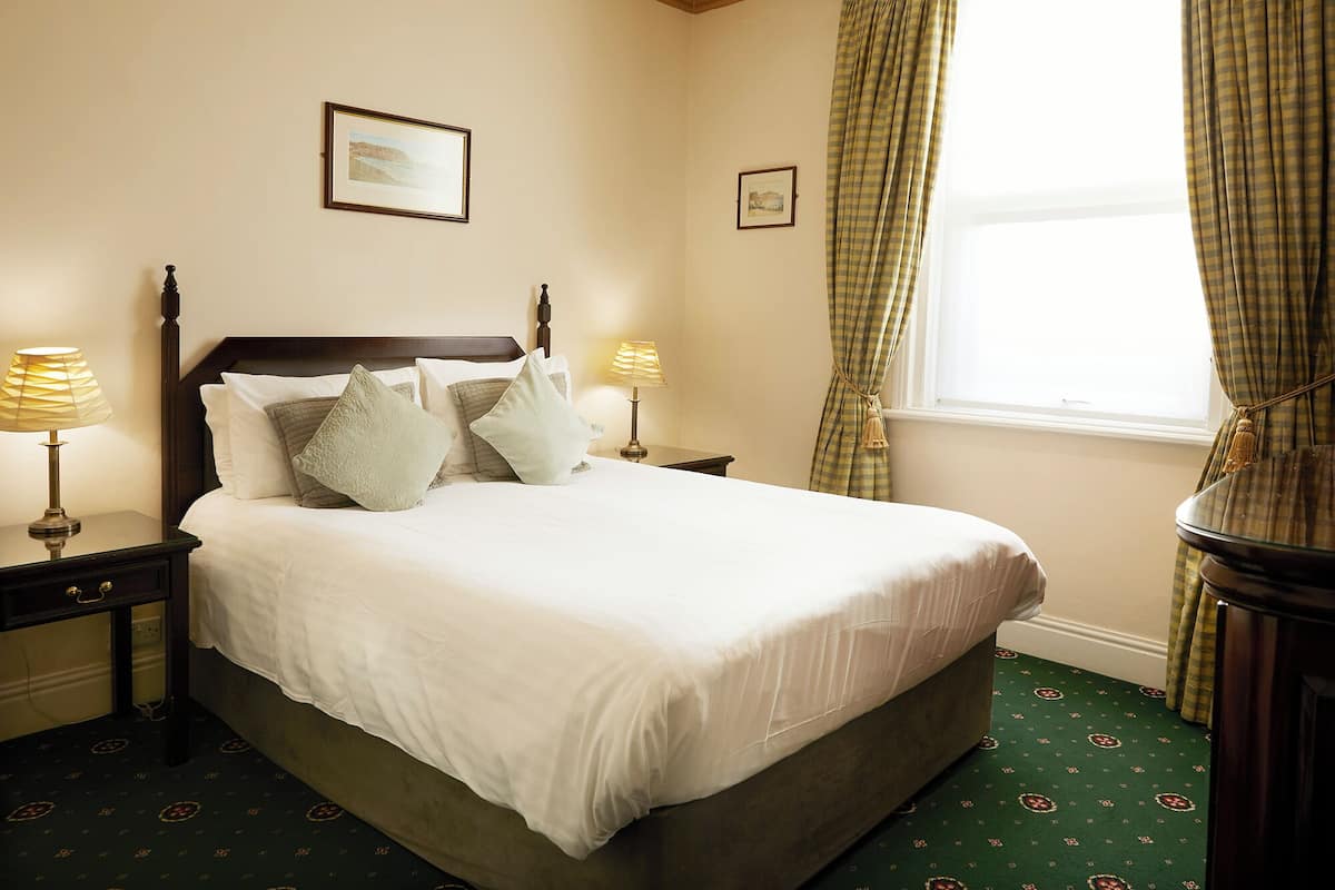 A neatly made bed with white linens and multiple pillows, a wooden headboard, two bedside lamps, and a window with curtains.