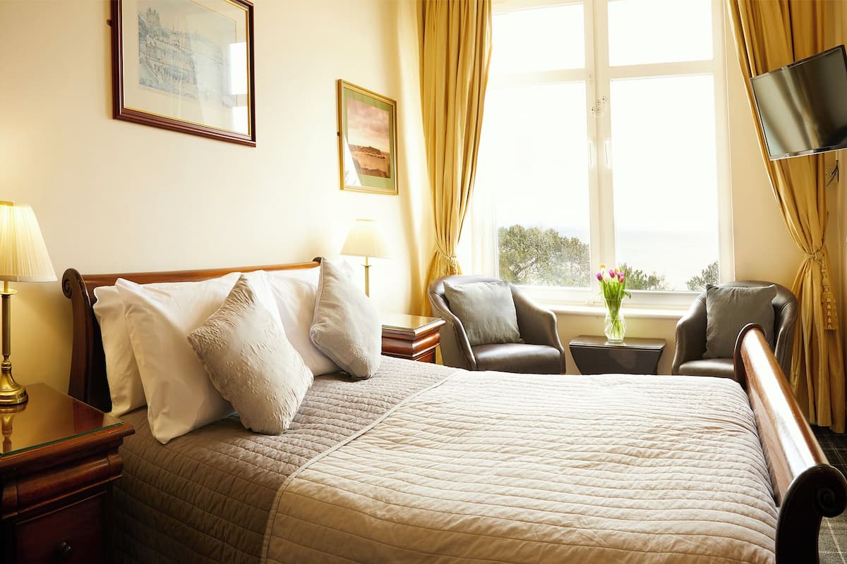 A hotel room with a large bed, two chairs, a small table with a vase of flowers, and a television on a stand.