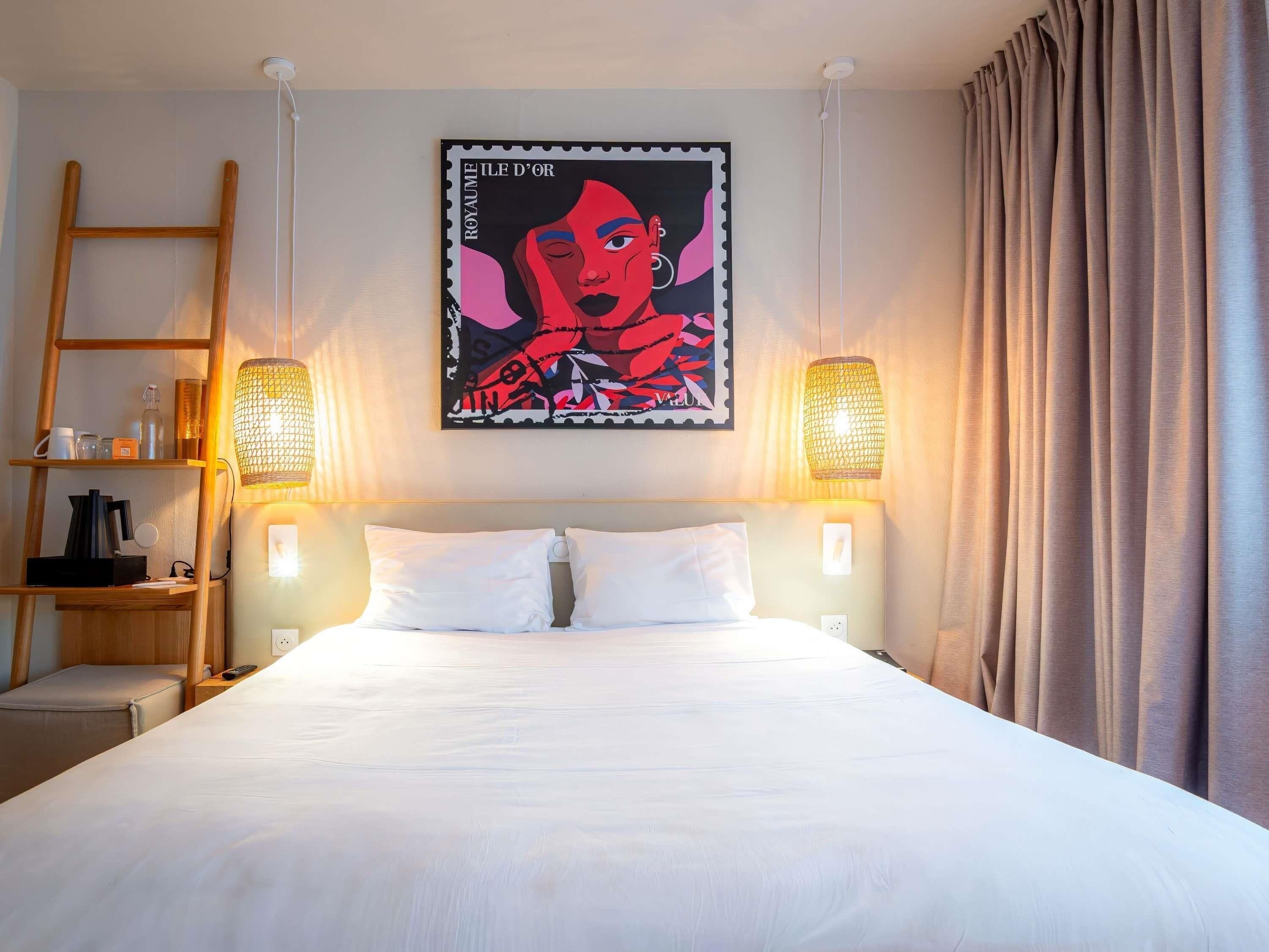 A hotel room with a bed, two bedside lamps, a shelf with toiletries, a ladder, and a wall-mounted artwork.