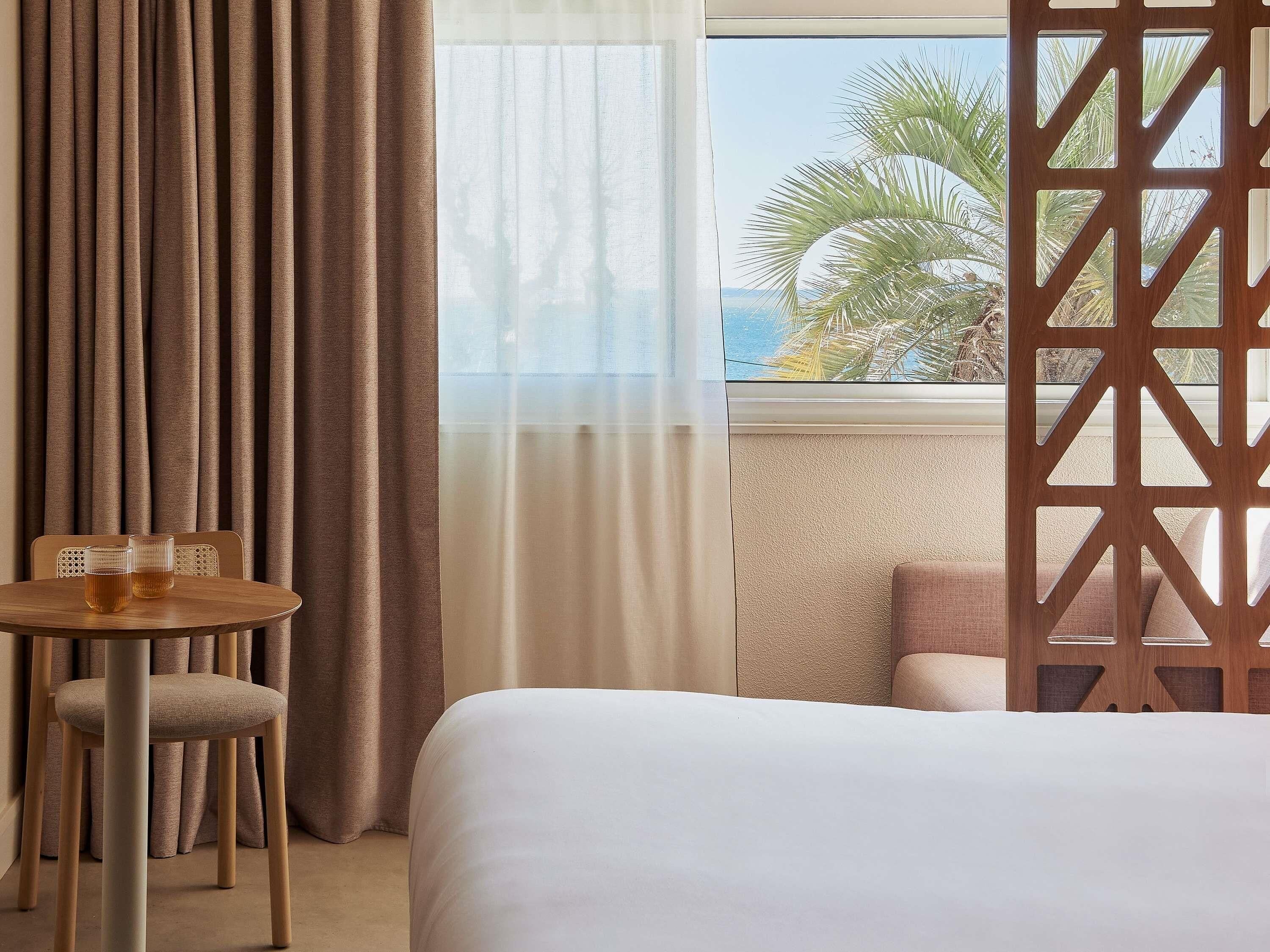 Une chambre d’hôtel avec un lit, une petite table sur laquelle sont posés deux verres, une chaise et une vue sur des palmiers par la fenêtre.