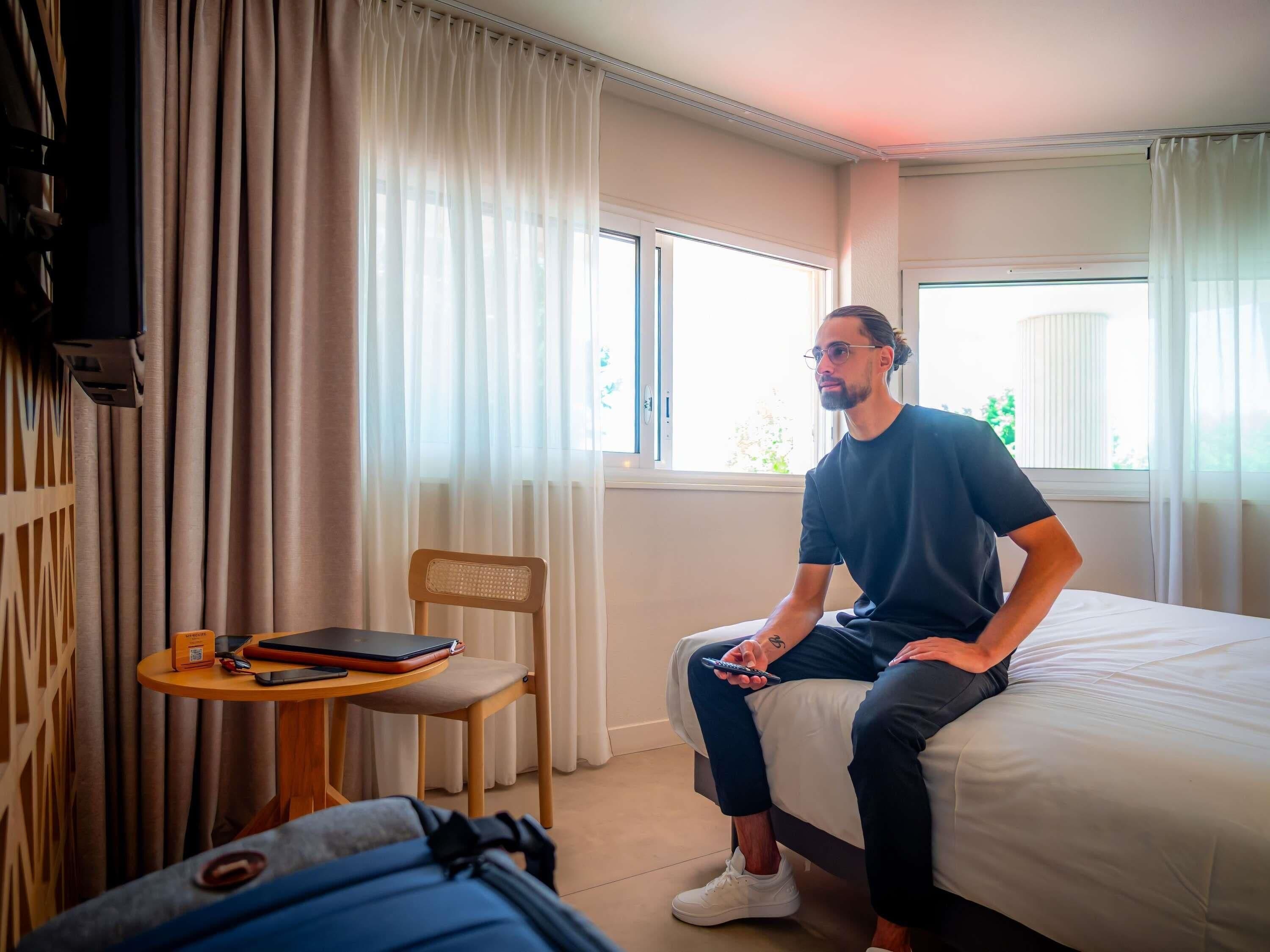 A hotel room with a bed, a chair, a small table with a laptop and a phone, and a suitcase on the floor.