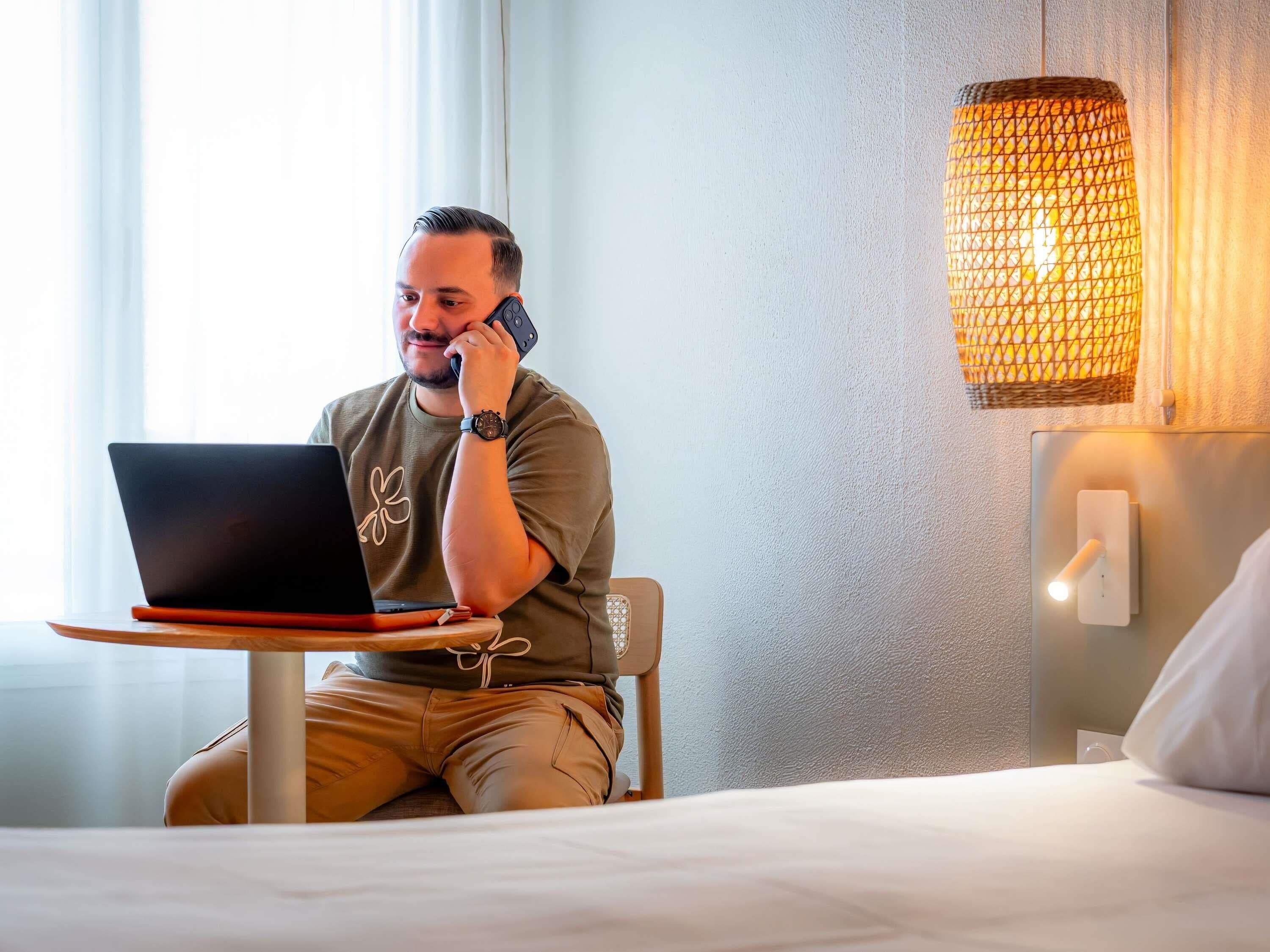 A person sitting at a table with a laptop, a phone, and a lamp in a room with a bed and a wall-mounted light fixture.