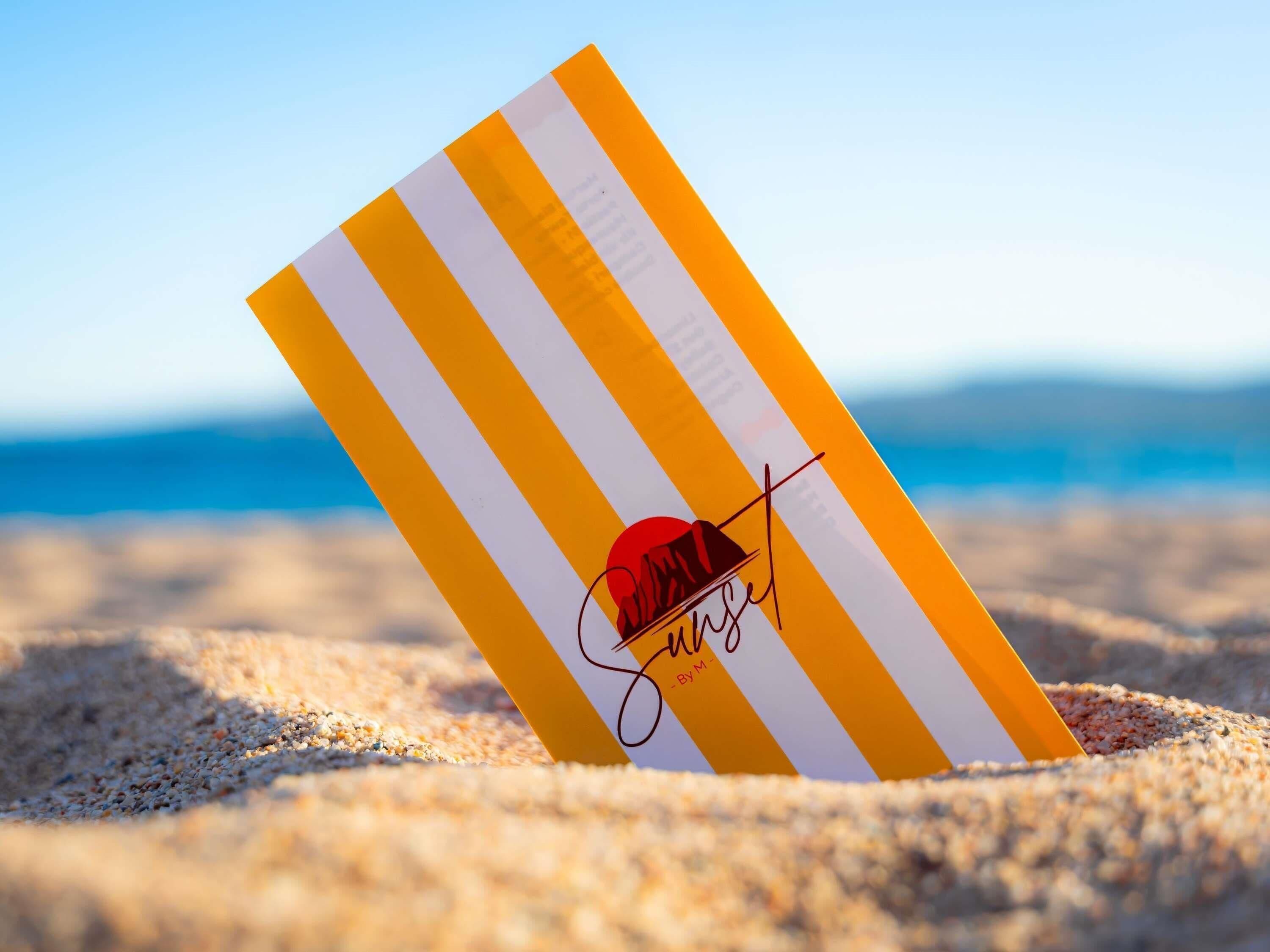 A striped beach towel with a sun hat and the word 'Sunrise' on a sandy beach.