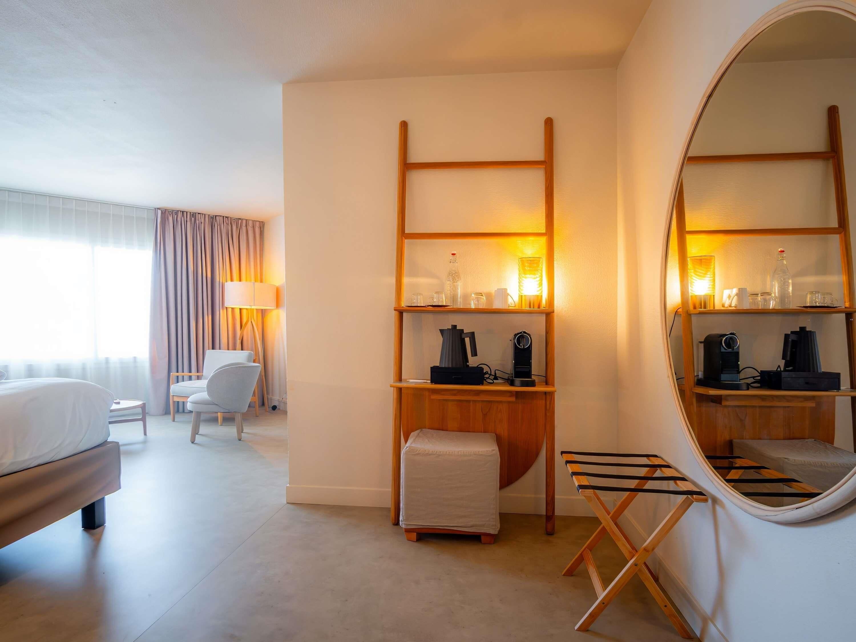 Habitación de hotel con cama, una silla, un escritorio con lámpara, espejo y estantes con objetos.