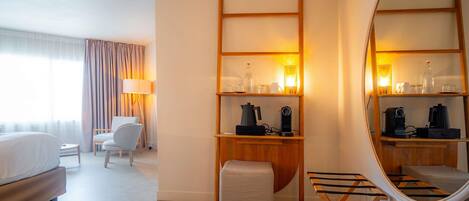 Habitación de hotel con cama, una silla, un escritorio con lámpara, espejo y estantes con objetos.
