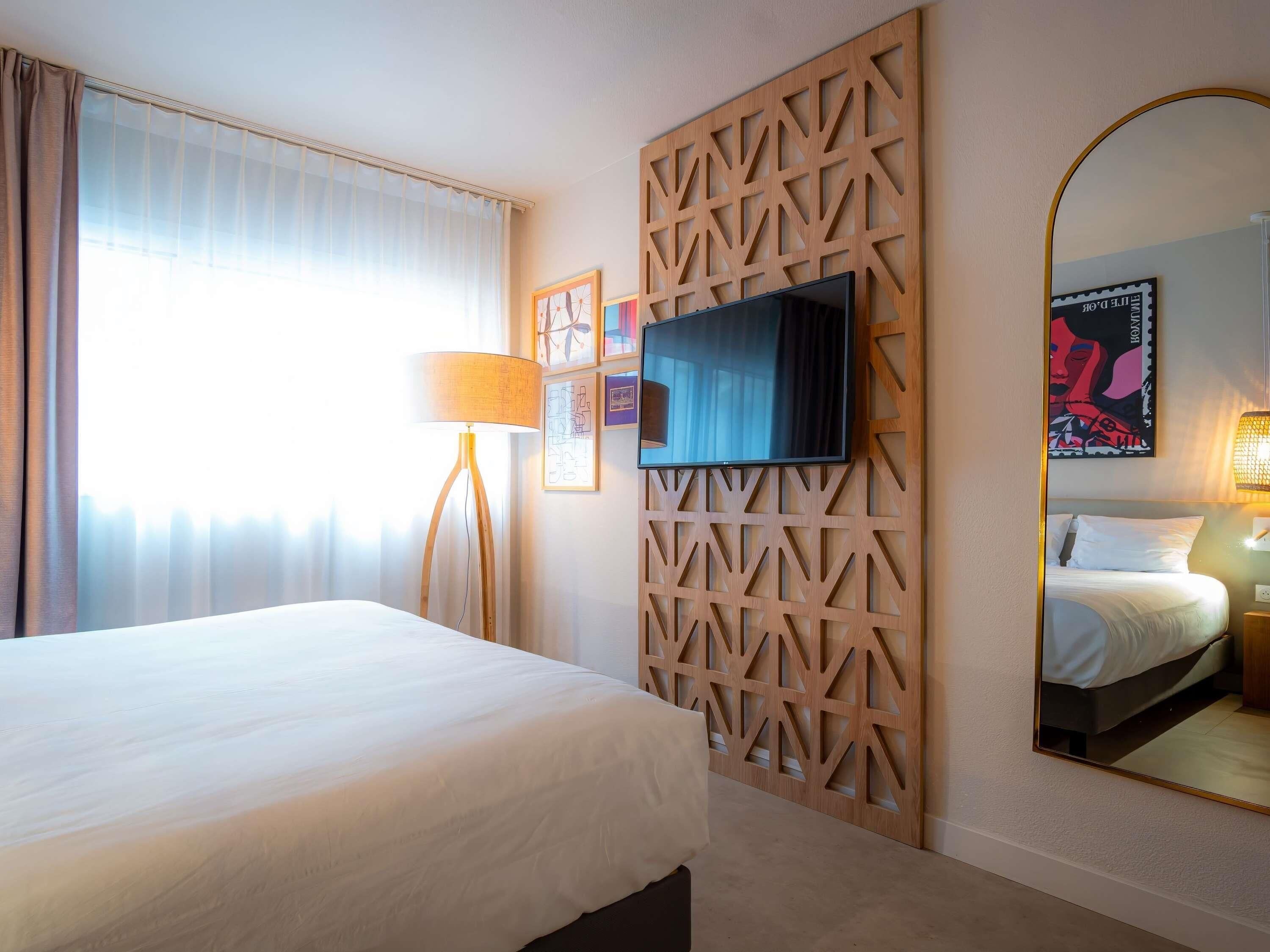 A hotel room with a bed, a TV mounted on a wooden panel, a large mirror, and a window with curtains.