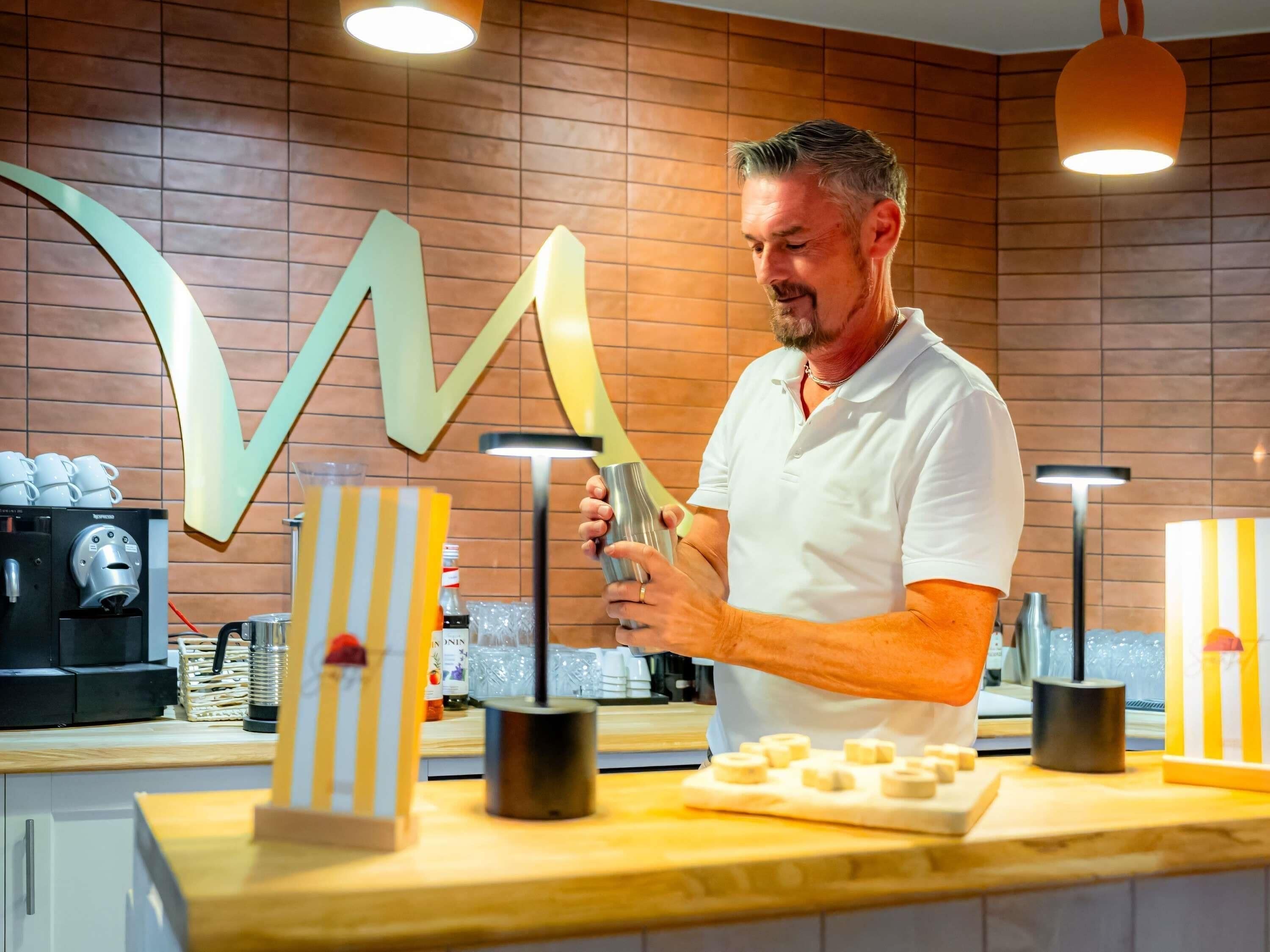 A person preparing food at a counter with a coffee machine, a large decorative letter, and a window in the background.