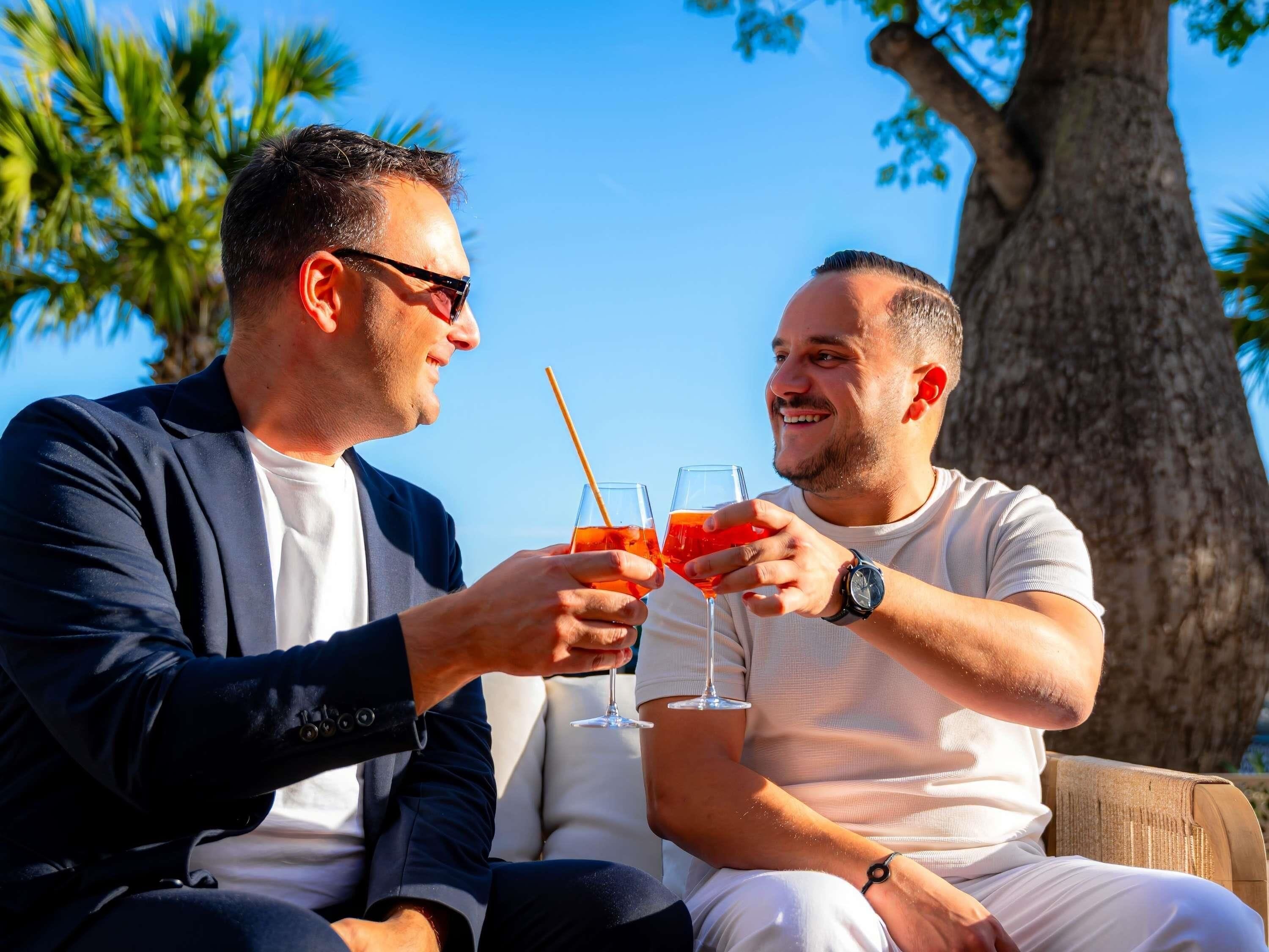 Dos personas sentadas al aire libre, sosteniendo vasos con bebidas, con un árbol y un cielo despejado al fondo.