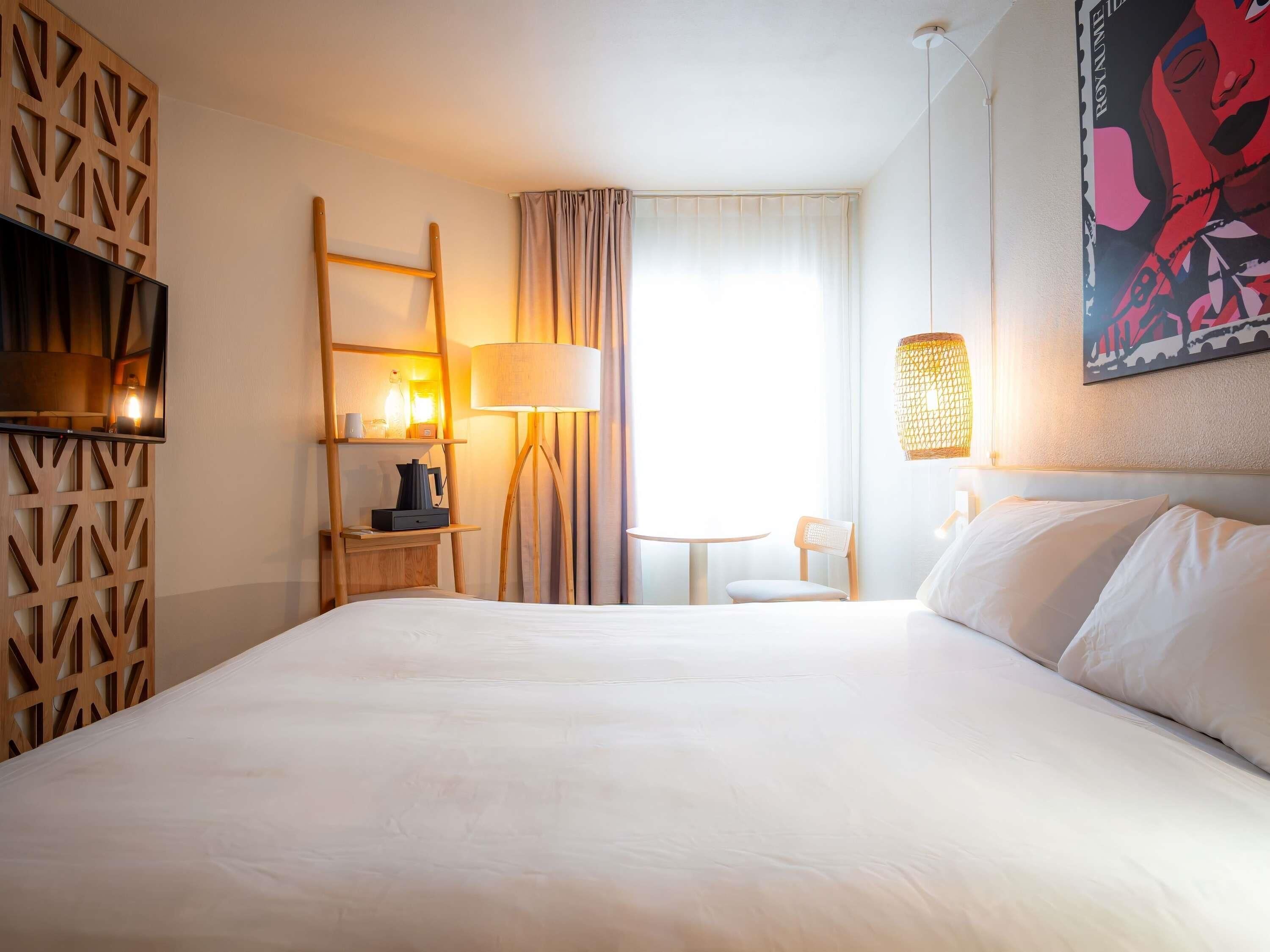 Habitación de hotel con una cama grande, un escritorio con lámpara, una silla, un televisor y una ventana con cortinas.