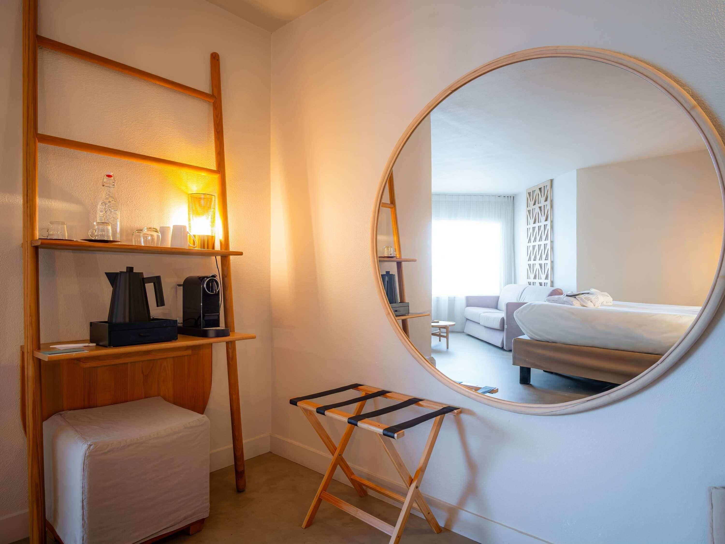 A hotel room with a bed, a small seating area, a coffee machine, a shelf with items, and a round mirror.