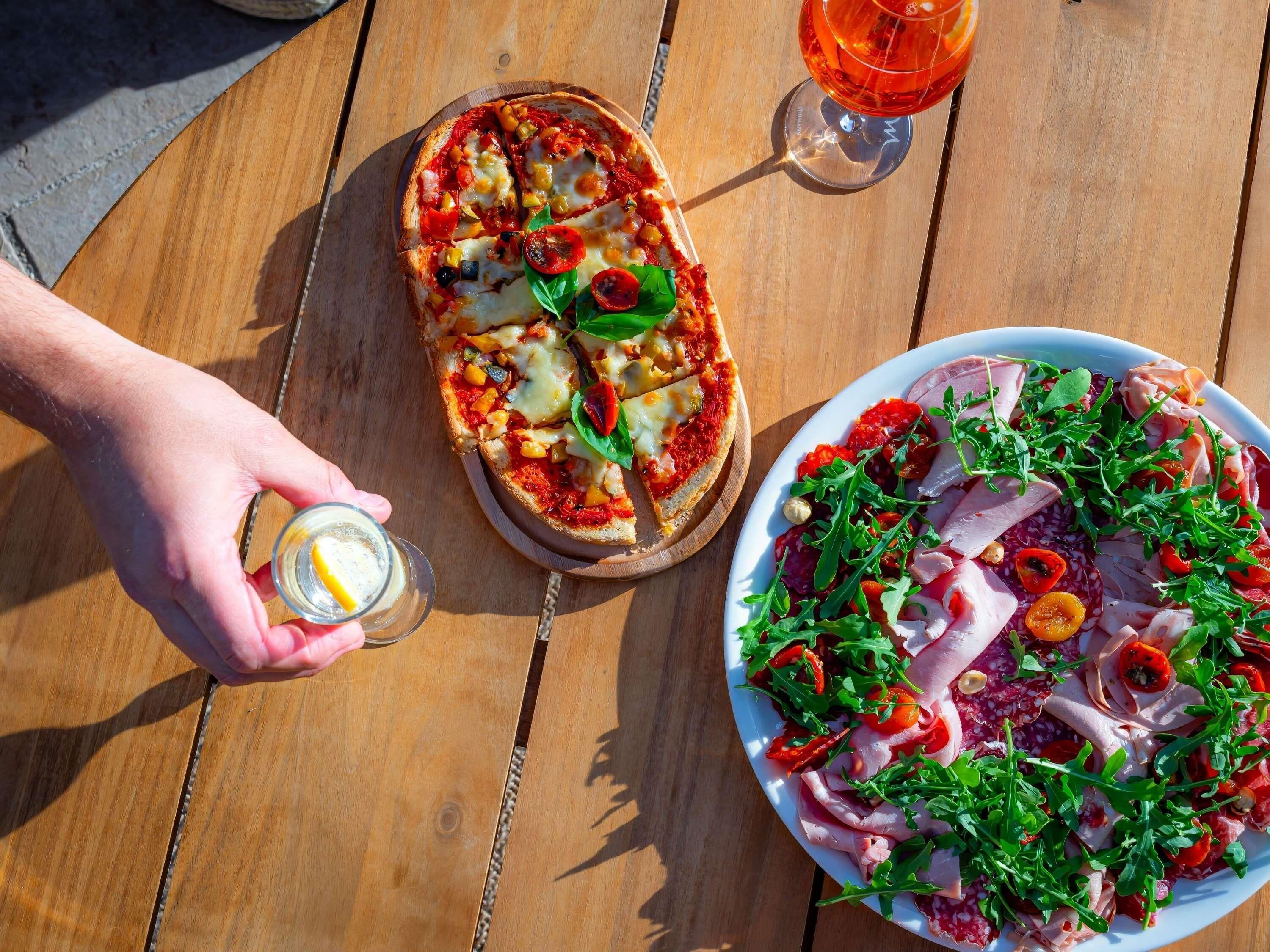 Eine Pizza auf einem Holzboden, eine Platte mit Wurstwaren und Gemüse sowie ein Glas Getränk auf einem Holztisch.