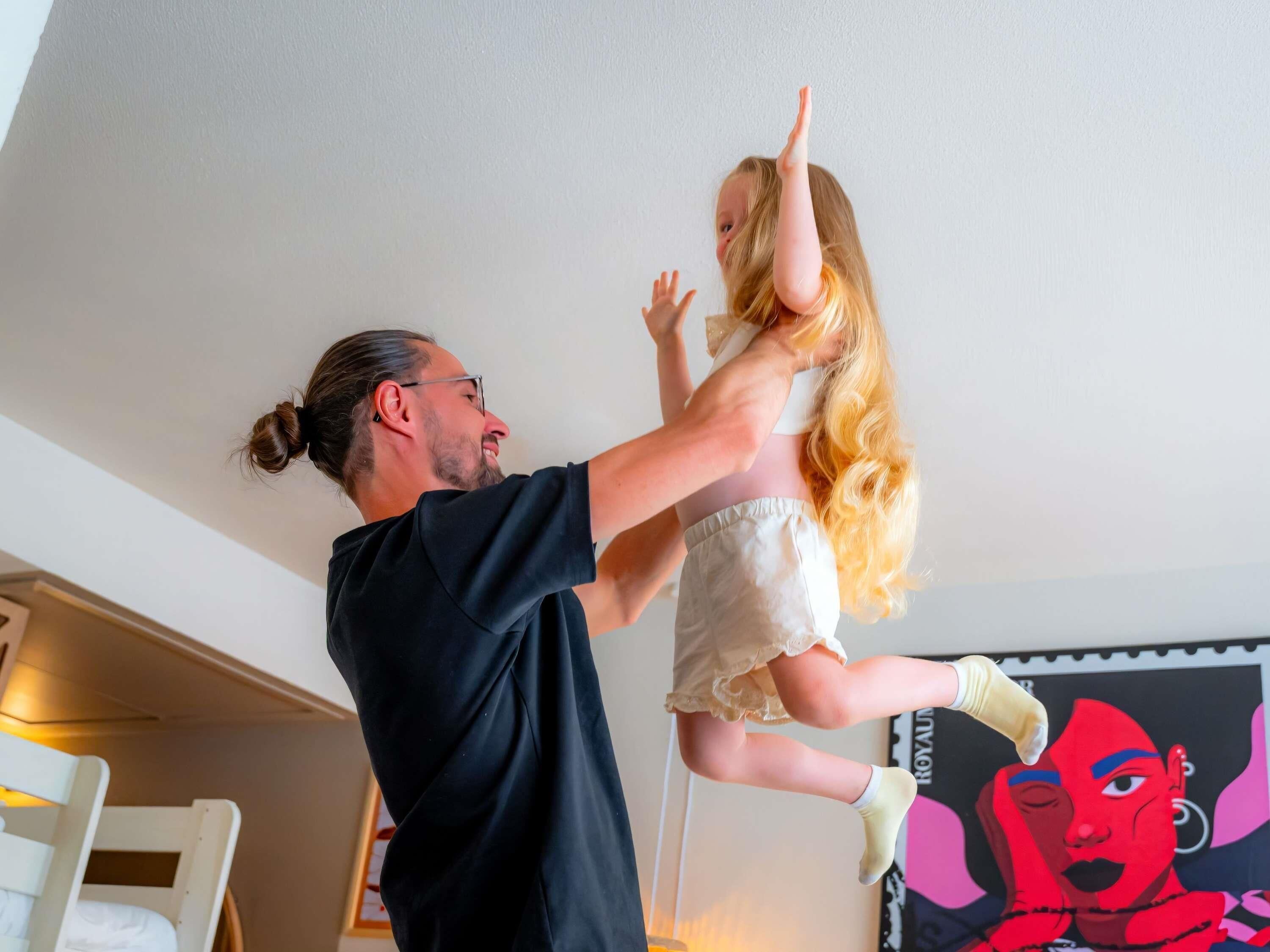 Eine Person hebt eine andere Person in einem Zimmer mit einem Bett und einer Wand mit einem Plakat.