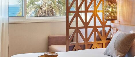 A hotel room with a bed, a view of palm trees and the sea, a wooden partition, and a woven lamp.