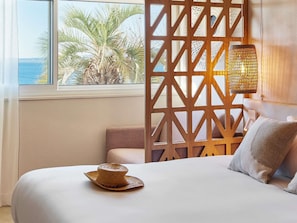Habitación de hotel con cama, vista a palmeras y al mar, una pared divisoria de madera y una lámpara de fibras naturales.