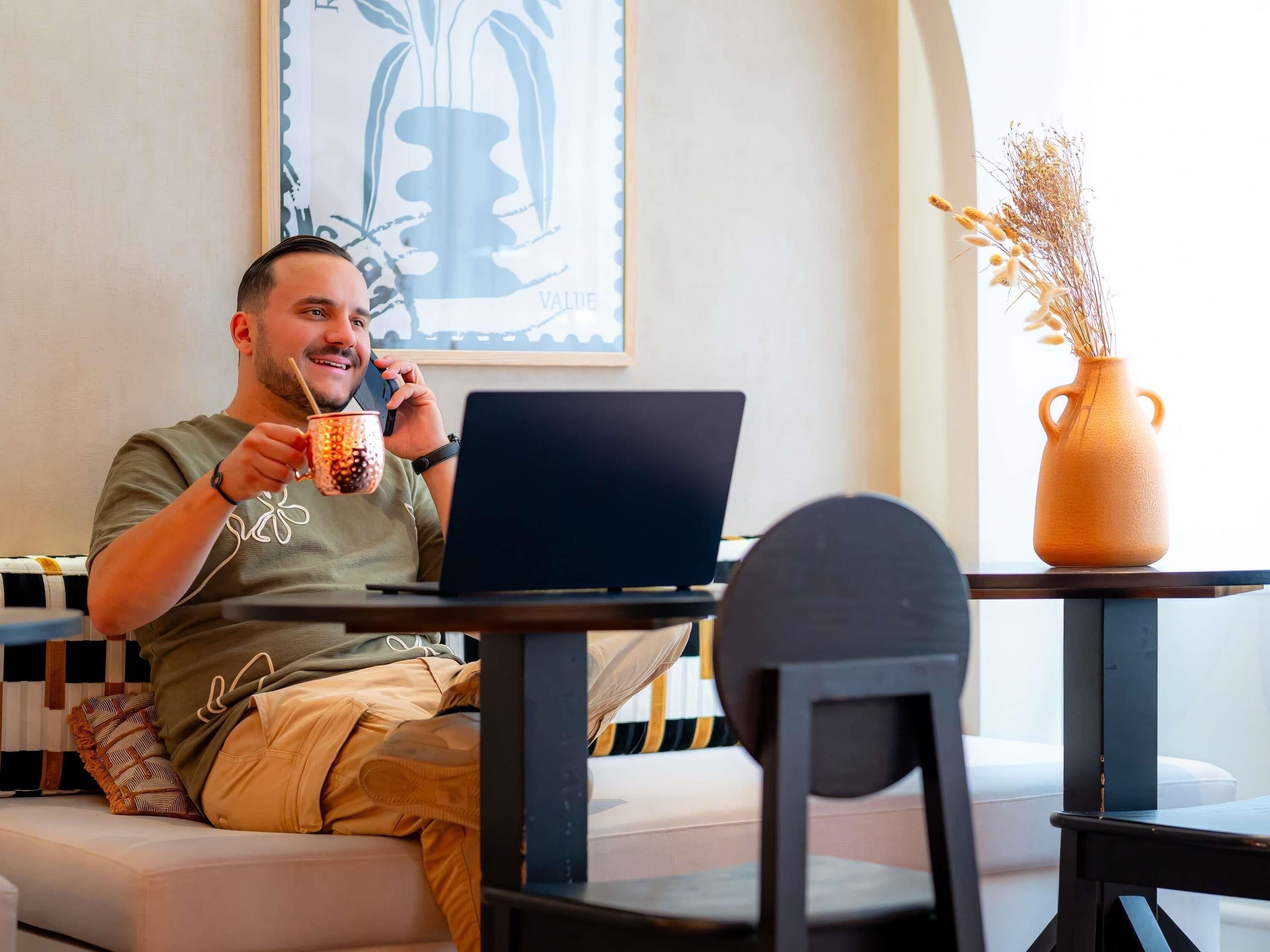 A person sitting on a sofa with a laptop, holding a mug, and a vase with dried plants on a table.