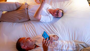 Dos personas recostadas en una cama con ropa blanca, una leyendo un libro y la otra usando un teléfono inteligente.