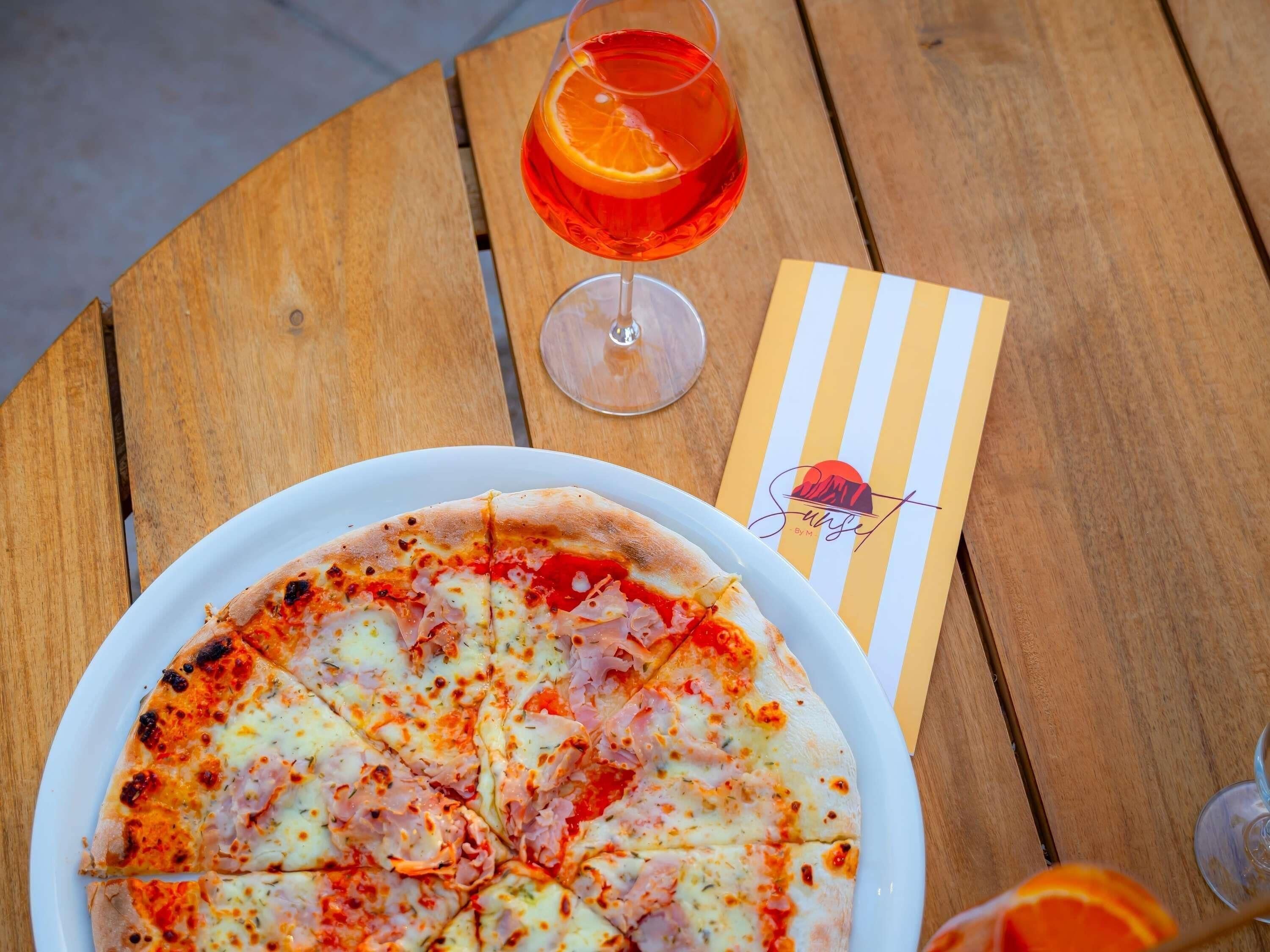 A pizza on a white plate, a glass of orange juice, and a striped napkin on a wooden table.
