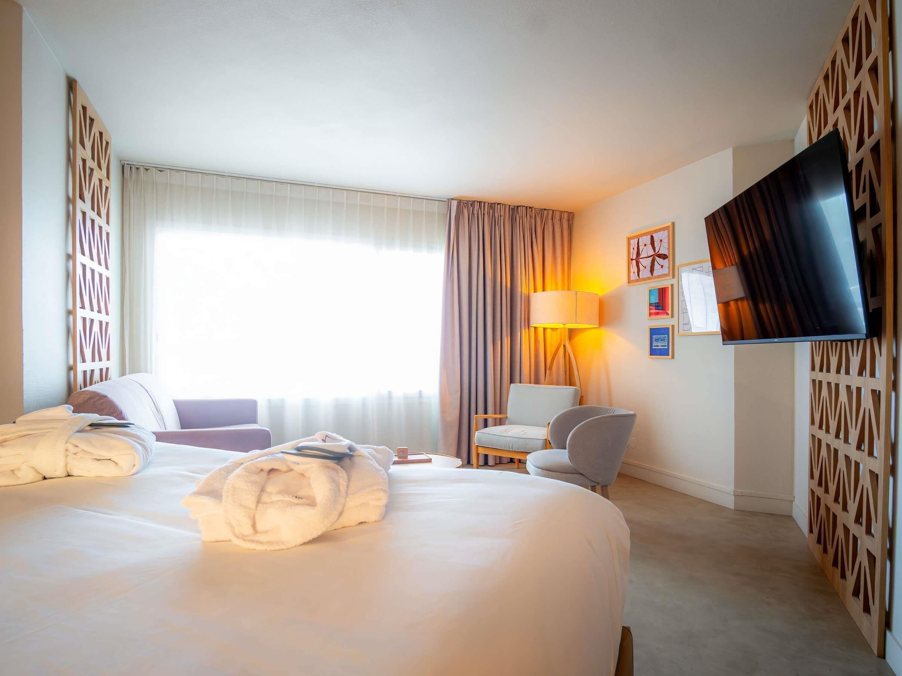 Une chambre d’hôtel avec un grand lit, un bureau, une chaise, une télévision et une fenêtre avec des rideaux.