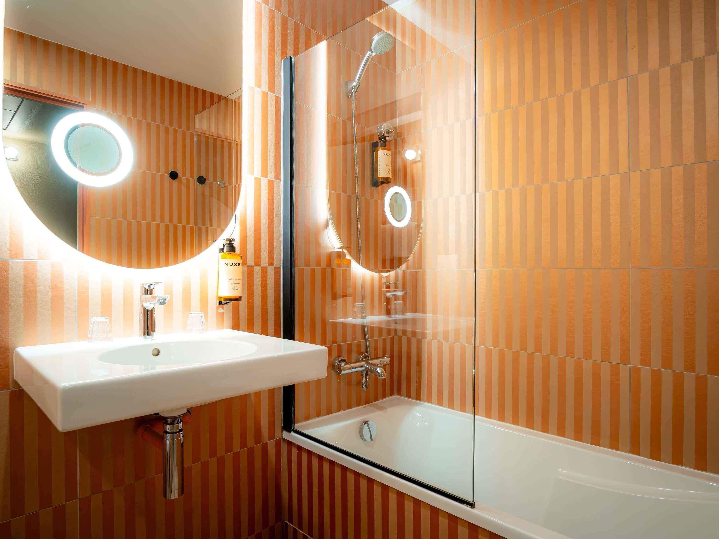 Ein Badezimmer mit einem weißen Waschbecken, einem Spiegel mit runder Beleuchtung, einer Duschkabine aus Glas und orangefarben gestreiften Wänden.
