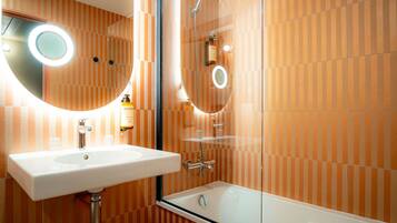 Ein Badezimmer mit einem weißen Waschbecken, einem Spiegel mit runder Beleuchtung, einer Duschkabine aus Glas und orangefarben gestreiften Wänden.