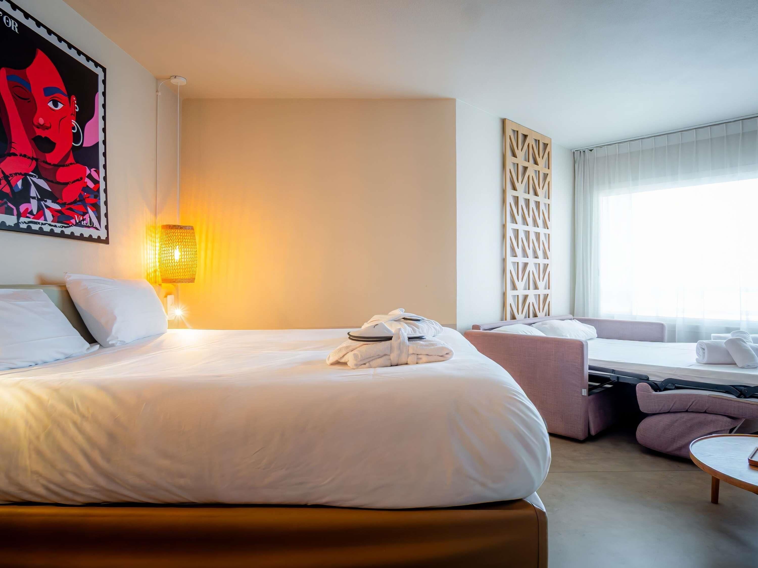 Habitació d'hotel amb un llit gran, una taula de treball, una cadira, una làmpada i una finestra amb cortines.