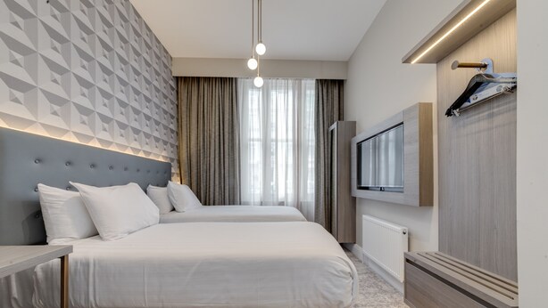 A modern hotel room with two beds, a flat-screen TV, a wooden headboard, a radiator, and a patterned wall. - Mornington Victor Hotel London Belgravia (London)