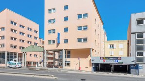 Un edificio de varios pisos con fachada plana, varias ventanas y un cartel que dice 'P'tayant Médicis' al nivel de la calle.