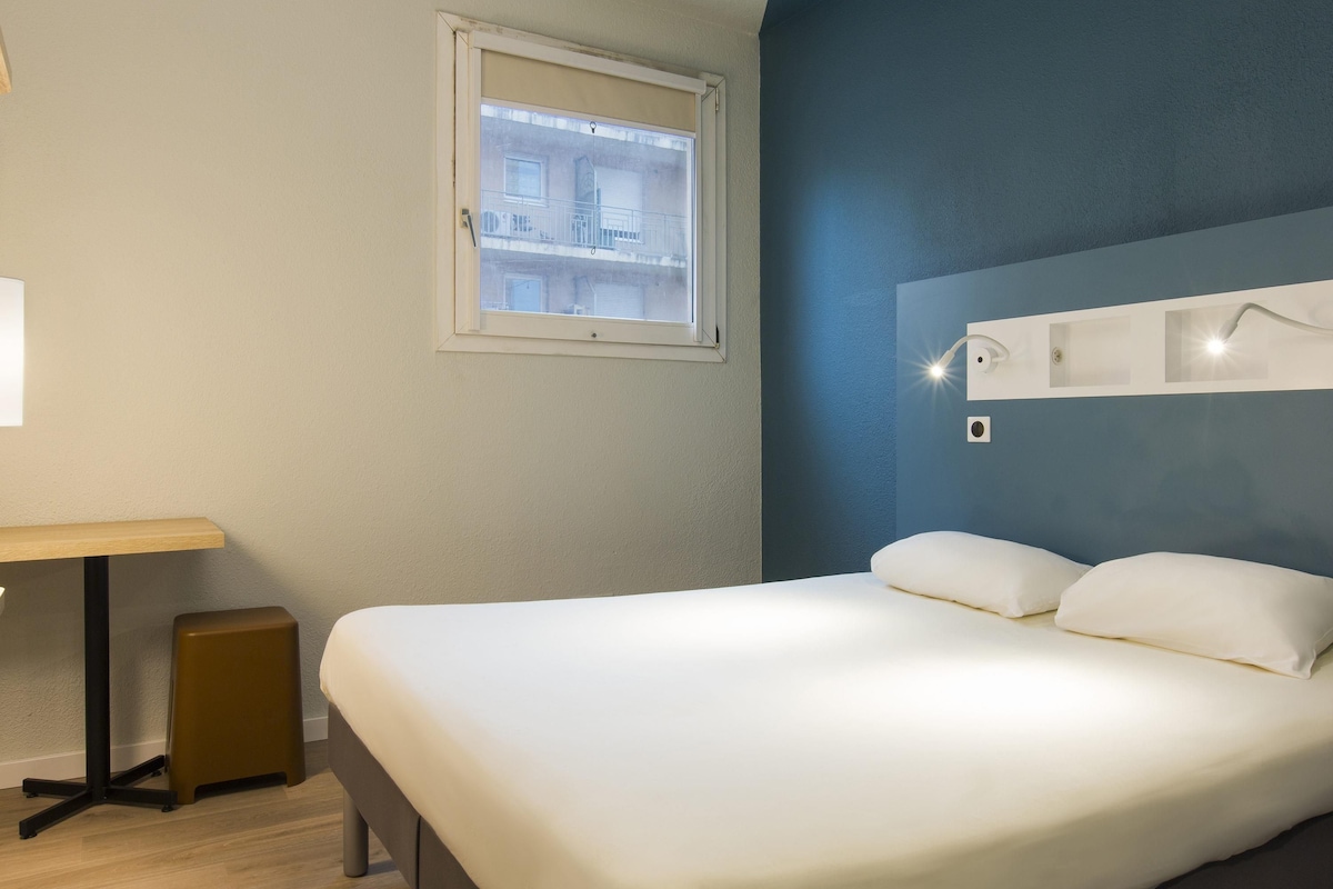 A hotel room with a bed, a desk, a small stool, a window, and a wall-mounted light fixture.