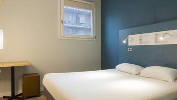 A hotel room with a bed, a desk, a small stool, a window, and a wall-mounted light fixture.