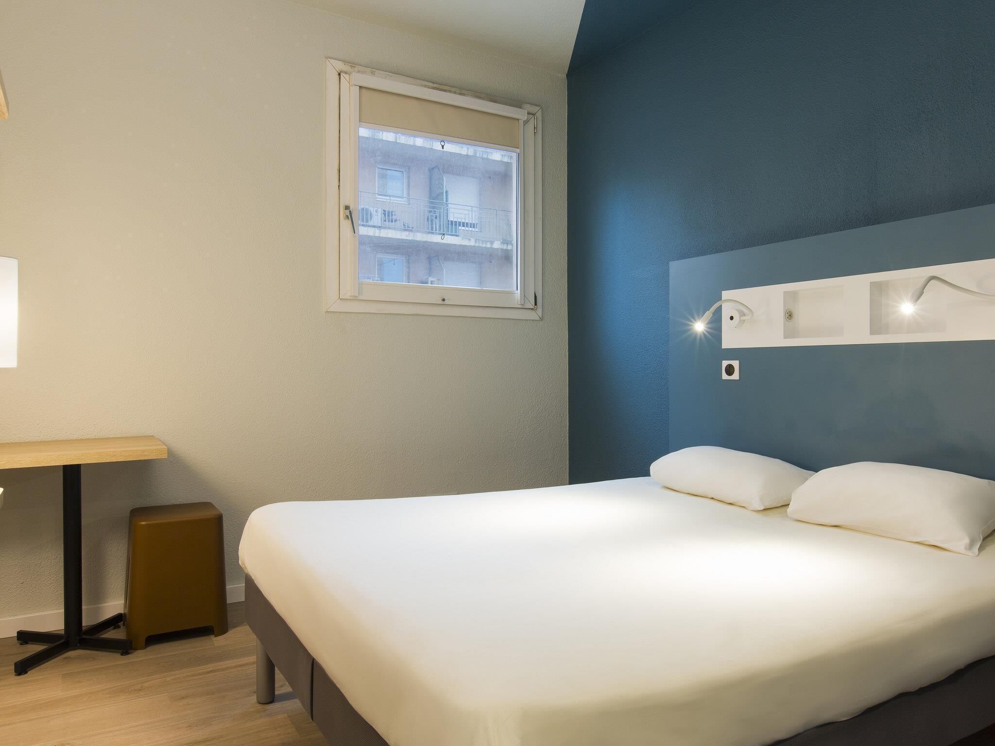 Una habitación de hotel con cama, escritorio, ventana y una lámpara fijada a la pared.