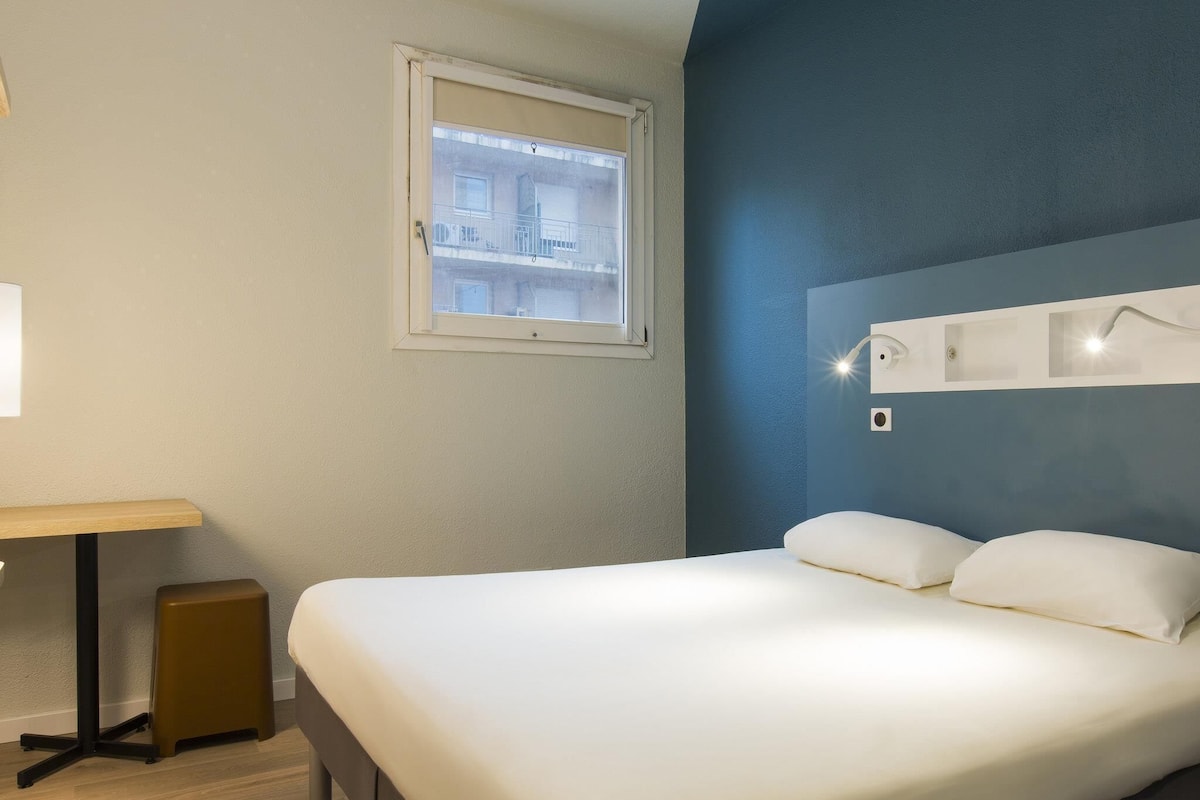 Una habitación de hotel con cama, escritorio, ventana y una lámpara fijada a la pared.