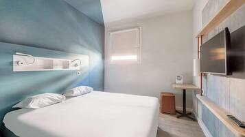 A modern hotel room with a bed, a TV mounted on the wall, a small table, and a window.