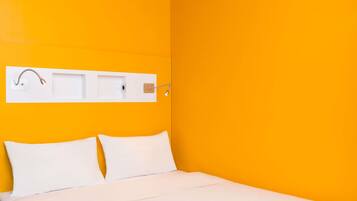 Una habitación con cama, ropa de cama blanca, un estante blanco montado en la pared con una toalla y un gancho, y una lámpara en el techo.